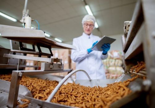 Close up view of a salt stick snacks being processed through a production line in food factory while being inspected by a female young worker with a tablet in a sterile cloth.